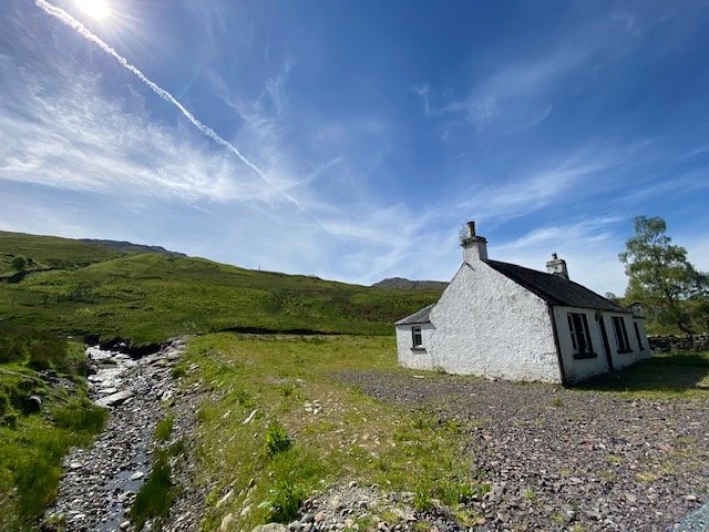 A white cottage beside a stream in a wide green glen