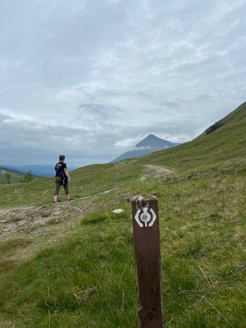 A walker on the trail beside a waymarker post, with mountains ahead